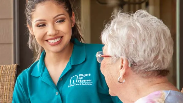 A caregiver in turquoise scrubs shares a warm moment with an elderly female client wearing glasses.