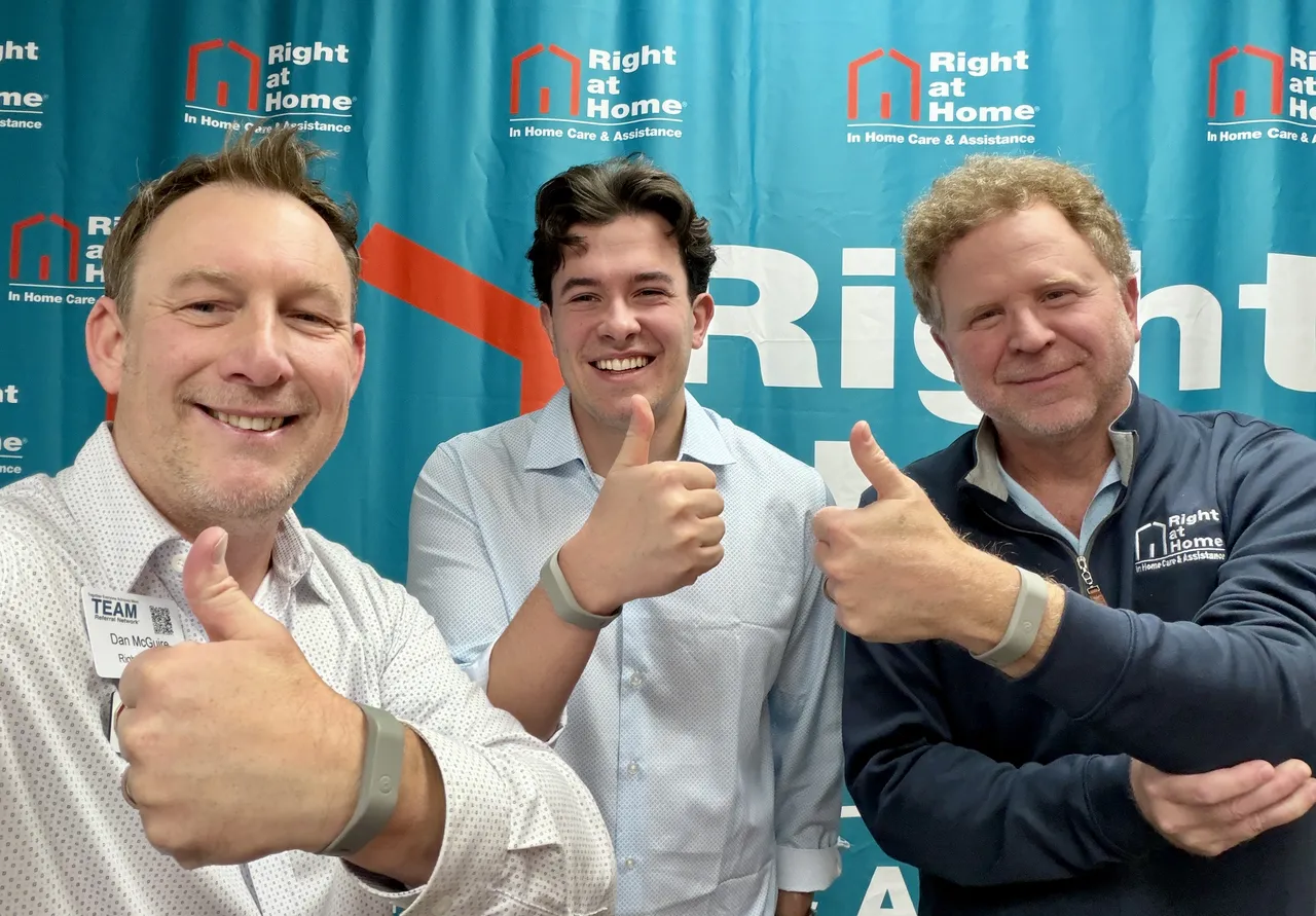 Three employees stand in front of a Right at Home company backdrop, giving thumbs-up in a positive pose.