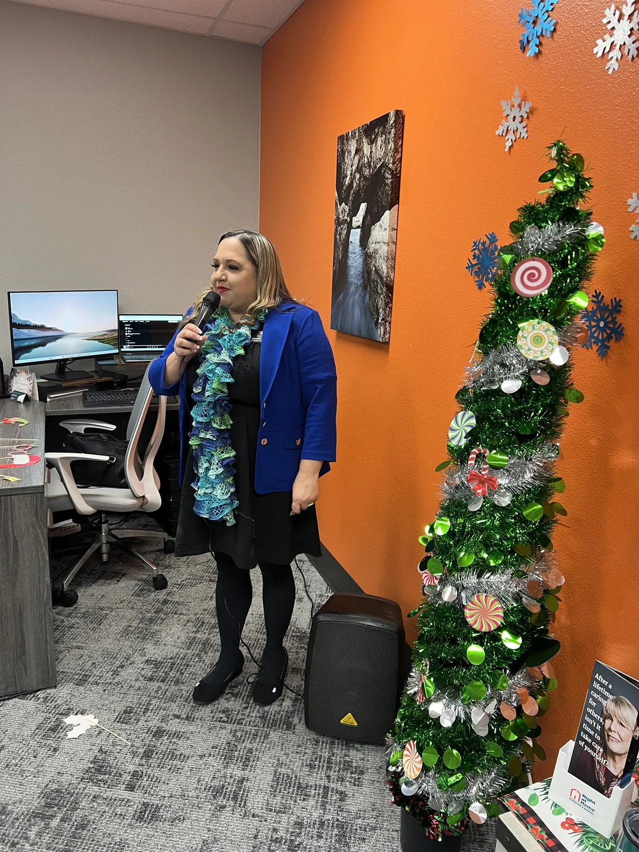 A woman in a blue jacket sits on a chair in an indoor space with orange walls, with green and silver Christmas wreath decorations hanging above her.