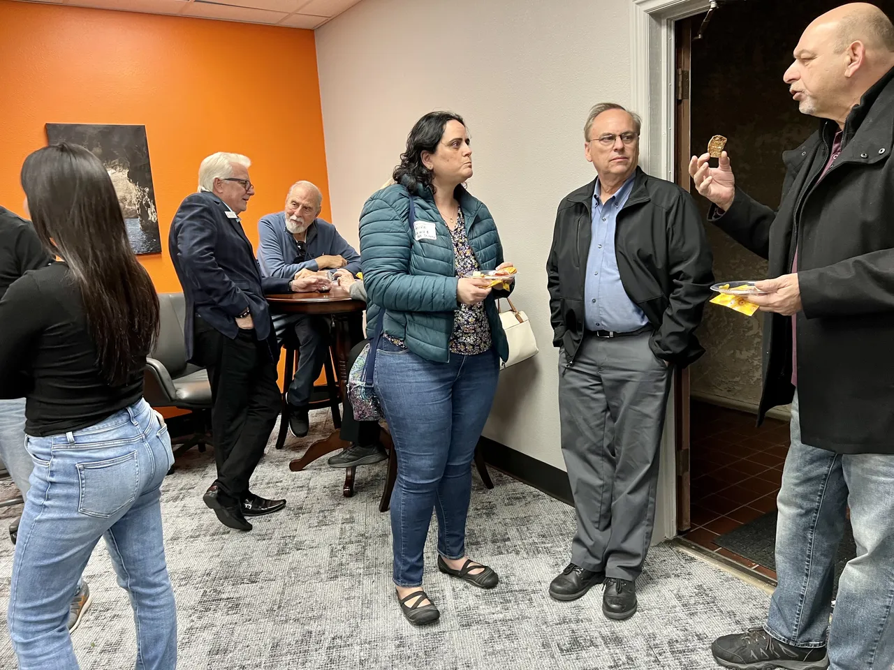 Several people gather in an indoor space with orange walls, holding food and drinks.
